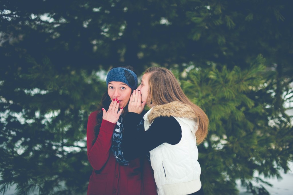Two women sharing secrets, outside and in front of trees.